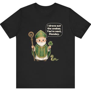 Puede incluir: Una camiseta negra con una ilustraci&oacute;n de dibujos animados de un obispo sosteniendo un bast&oacute;n de madera y un tr&eacute;bol verde. Una peque&ntilde;a serpiente verde est&aacute; en el suelo. Un globo de texto contiene: I drove out the snakes. You're next, Monday.