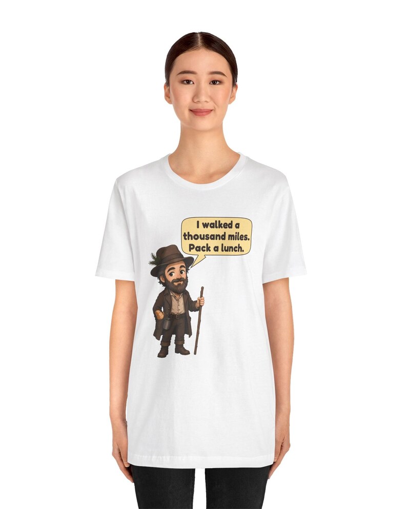 May include: A white short-sleeve t-shirt featuring a cartoon illustration of a bearded person wearing a brown hat and coat, holding a walking stick. A speech bubble above the character contains the text: I walked a thousand miles. Pack a lunch.