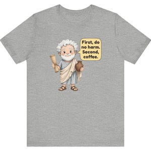 May include: A heather gray t-shirt featuring a cartoon illustration of an elderly person with white hair and a beard, wearing a light blue tunic and sandals. The figure holds a scroll and a brown pot, with a speech bubble stating: First, do no harm. Second, coffee.