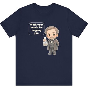 May include: A navy blue t-shirt featuring a cartoon illustration of a man in a suit holding a glass flask. A speech bubble next to the character contains the text: Wash your hands. I'm begging you. The design is centered on the front of the shirt.