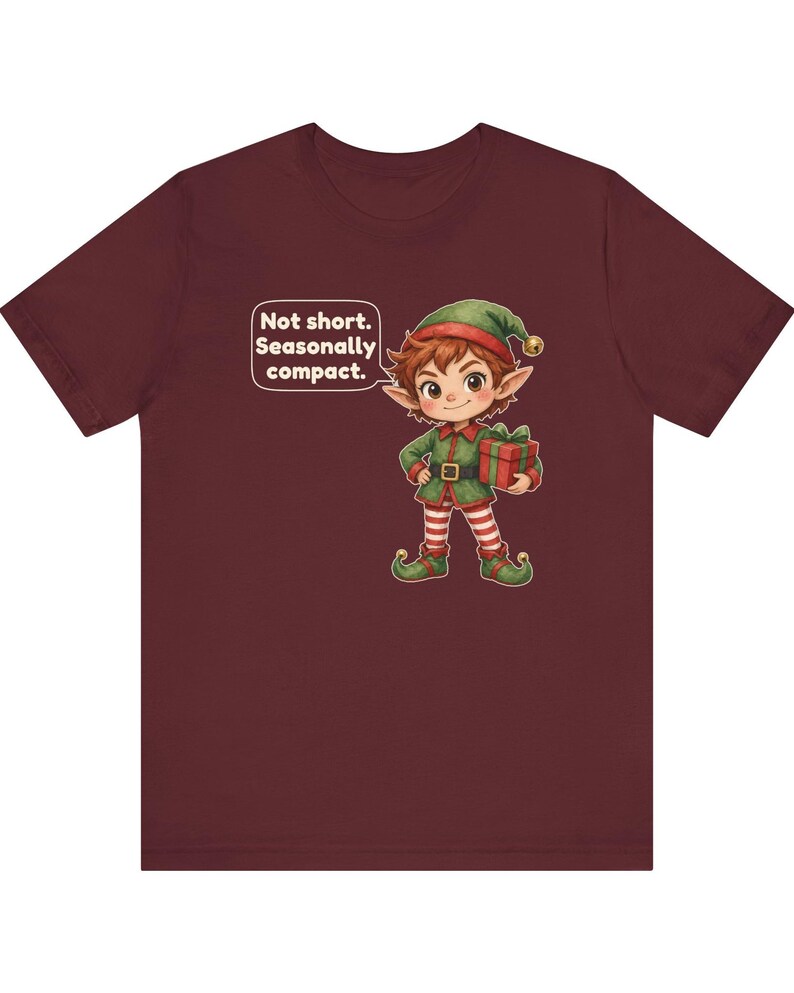 May include: A maroon short-sleeve t-shirt featuring a graphic of an elf wearing a green outfit and a hat with a bell, holding a gift. A speech bubble next to the elf contains the text: Not short. Seasonally compact.