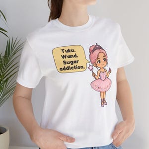 May include: A white t-shirt featuring a graphic of a cartoon ballerina with pink hair, a tiara, and a pink tutu, holding a star wand. A yellow speech bubble next to the character contains the text: Tutu. Wand. Sugar addiction.