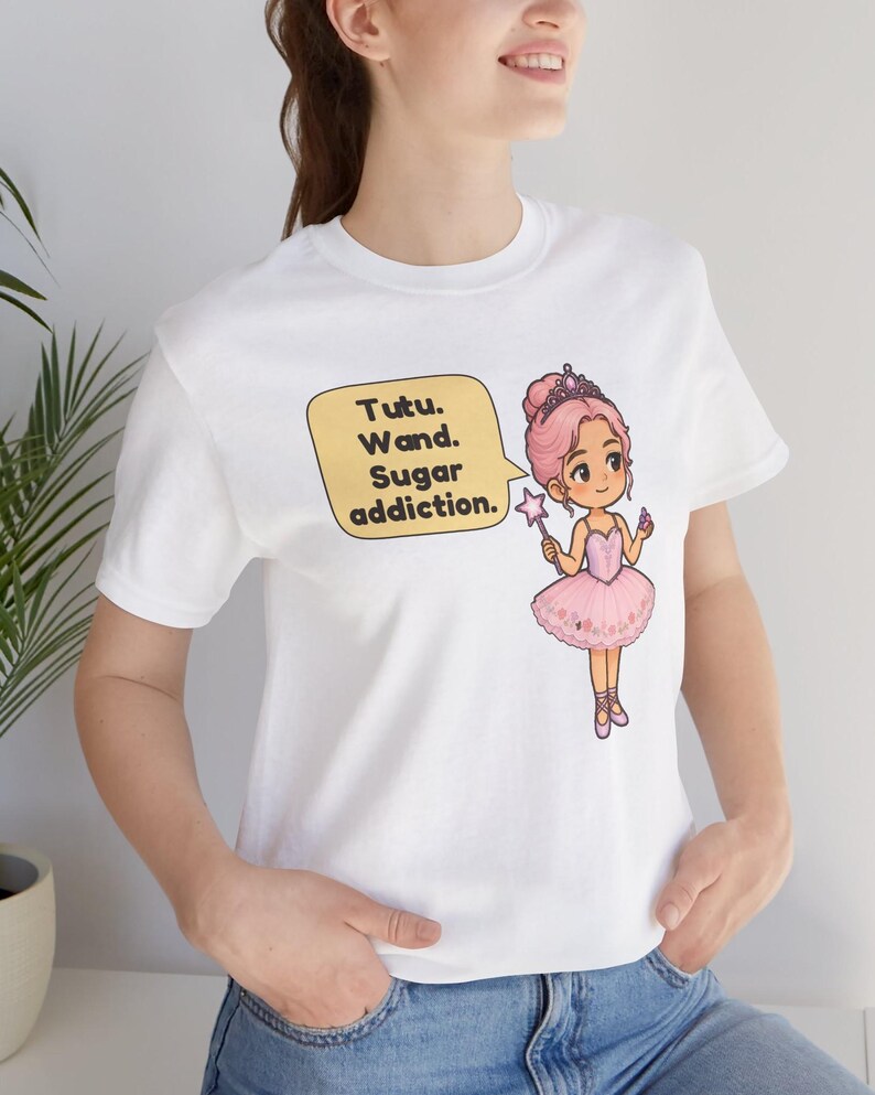 May include: A white t-shirt featuring a graphic of a cartoon ballerina with pink hair, a tiara, and a pink tutu, holding a star wand. A yellow speech bubble next to the character contains the text: Tutu. Wand. Sugar addiction.