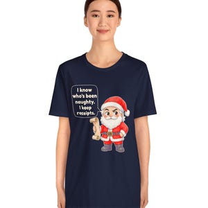 May include: A navy blue short-sleeve t-shirt featuring a graphic of Santa Claus holding a scroll. A speech bubble next to Santa contains the text: I know who's been naughty. I keep receipts. The design is centered on the front of the shirt.