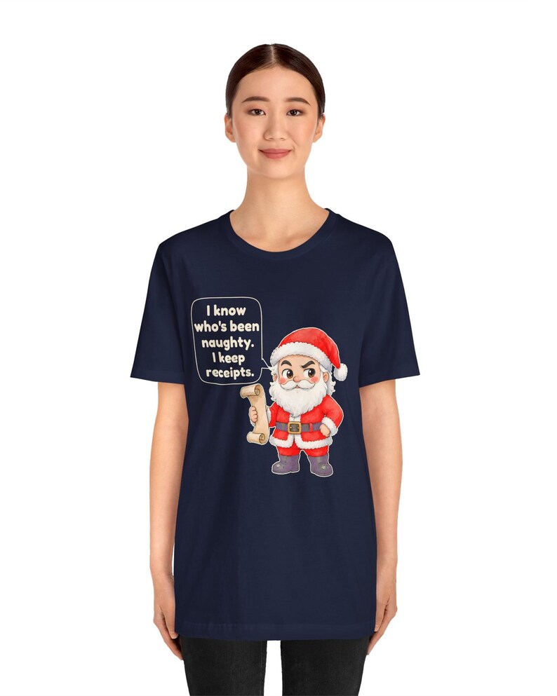 May include: A navy blue short-sleeve t-shirt featuring a graphic of Santa Claus holding a scroll. A speech bubble next to Santa contains the text: I know who's been naughty. I keep receipts. The design is centered on the front of the shirt.
