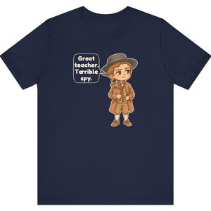 May include: A navy blue short-sleeve t-shirt featuring a cartoon illustration of a person in 18th-century attire, including a brown coat, hat, and breeches. A speech bubble next to the character contains the text: Great teacher. Terrible spy.