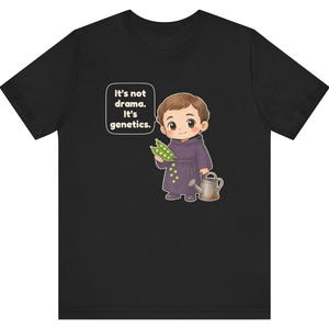 May include: A black t-shirt featuring a cartoon illustration of a monk holding a pea pod and a small watering can. A speech bubble next to the character contains the white text: It's not drama. It's genetics. The design is centered on the chest.