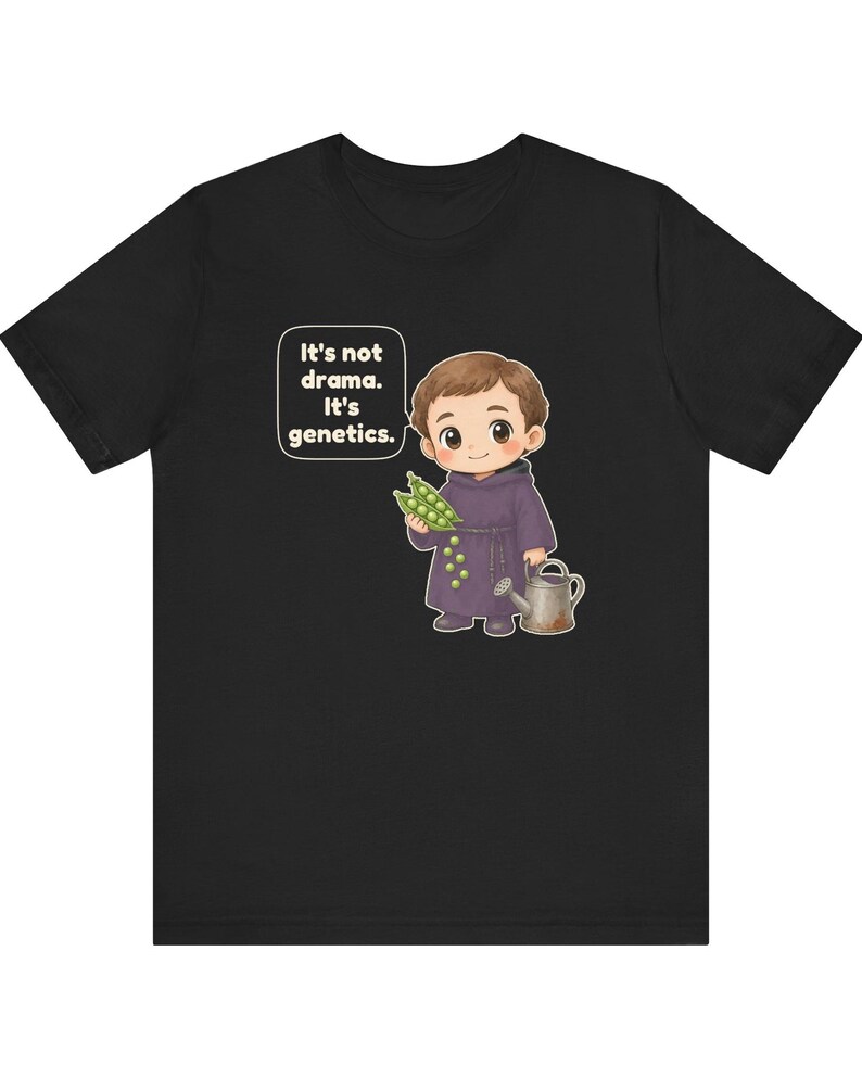 May include: A black t-shirt featuring a cartoon illustration of a monk holding a pea pod and a small watering can. A speech bubble next to the character contains the white text: It's not drama. It's genetics. The design is centered on the chest.