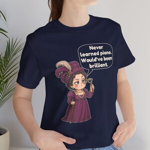 May include: A navy blue t-shirt featuring a cartoon illustration of a person in a purple Victorian-style dress and hat, holding a monocle. A speech bubble above contains the text: Never learned piano. Would've been brilliant.