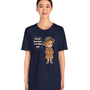 May include: A navy blue short-sleeve t-shirt featuring a cartoon illustration of a person wearing a brown hat, coat, and trousers. A speech bubble next to the character contains the text: Great teacher. Terrible spy.