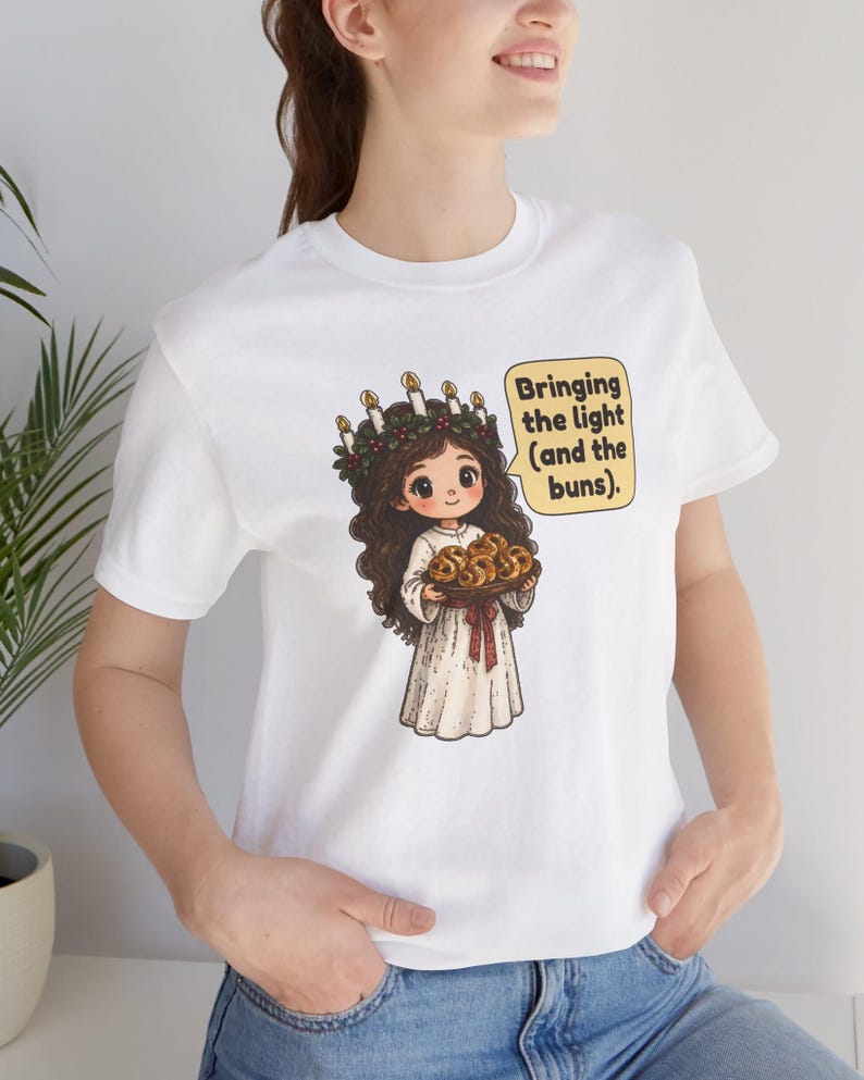 May include: A white t-shirt featuring a graphic of a character wearing a candle crown and holding a tray of buns. The character has long brown hair and wears a white robe. A speech bubble next to the character contains the text: Bringing the light (and the buns).