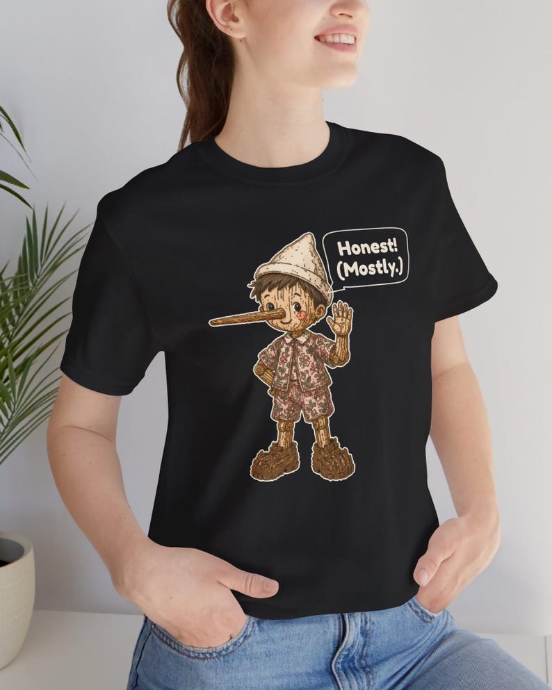 K&ouml;nnte beinhalten: Ein schwarzes Kurzarm-T-Shirt mit einer Grafik einer h&ouml;lzernen Puppenfigur mit langer Nase, die einen Hut und gemusterte Kleidung tr&auml;gt. Eine Sprechblase neben der Figur enth&auml;lt den Text: Honest! (Mostly.) Das Design ist mittig platziert.