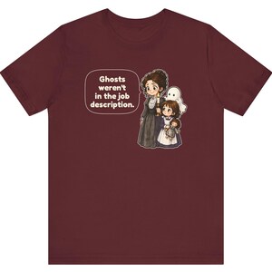 Op de afbeelding: Een bordeauxrood t-shirt met korte mouwen en een afbeelding van een persoon in een donkere jurk die een kaars vasthoudt, een kind en een klein spookje. Een tekstballon bevat de tekst: Ghosts weren't in the job description.