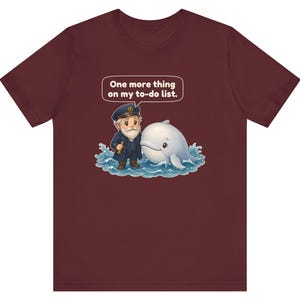 May include: A maroon short-sleeve t-shirt featuring a graphic of a cartoon sea captain and a white whale in blue water. A speech bubble above them contains the text, "One more thing on my to-do list." The design is centered on the chest.