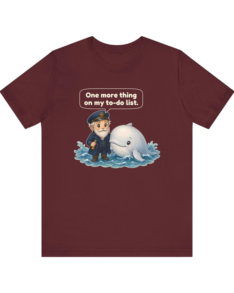 May include: A maroon short-sleeve t-shirt featuring a graphic of a cartoon sea captain and a white whale in blue water. A speech bubble above them contains the text, "One more thing on my to-do list." The design is centered on the chest.