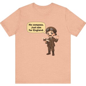May include: A peach-colored t-shirt featuring a cartoon illustration of a pilot with a mustache, goggles, and a brown suit holding a small airplane. A speech bubble above the character contains the text: No compass. Just aim for England.