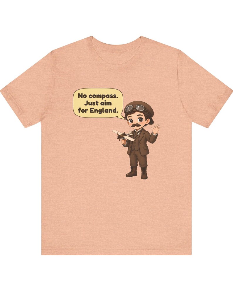 May include: A peach-colored t-shirt featuring a cartoon illustration of a pilot with a mustache, goggles, and a brown suit holding a small airplane. A speech bubble above the character contains the text: No compass. Just aim for England.