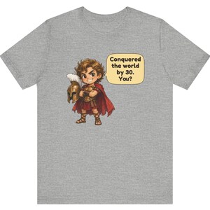 May include: A heather gray t-shirt featuring a cartoon illustration of a warrior in bronze armor and a red cape holding a helmet. A speech bubble next to the character contains the text: Conquered the world by 30. You?