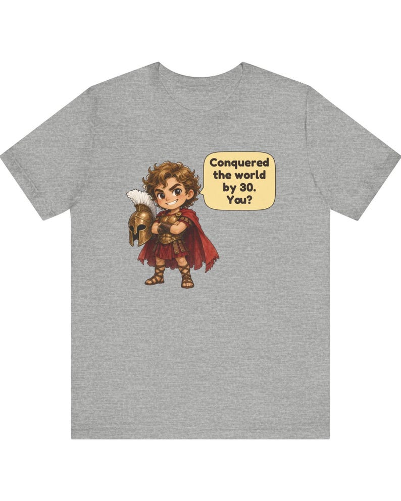 May include: A heather gray t-shirt featuring a cartoon illustration of a warrior in bronze armor and a red cape holding a helmet. A speech bubble next to the character contains the text: Conquered the world by 30. You?