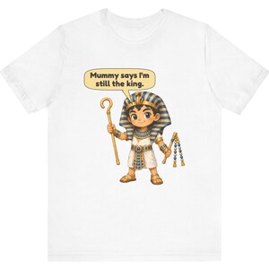 Może przedstawiać: Biały t-shirt z rysunkową ilustracją młodego faraona w niebiesko-złotym nakryciu głowy, trzymającego złote berło i bicz. Dymek nad postacią zawiera tekst, Mummy says I'm still the king.
