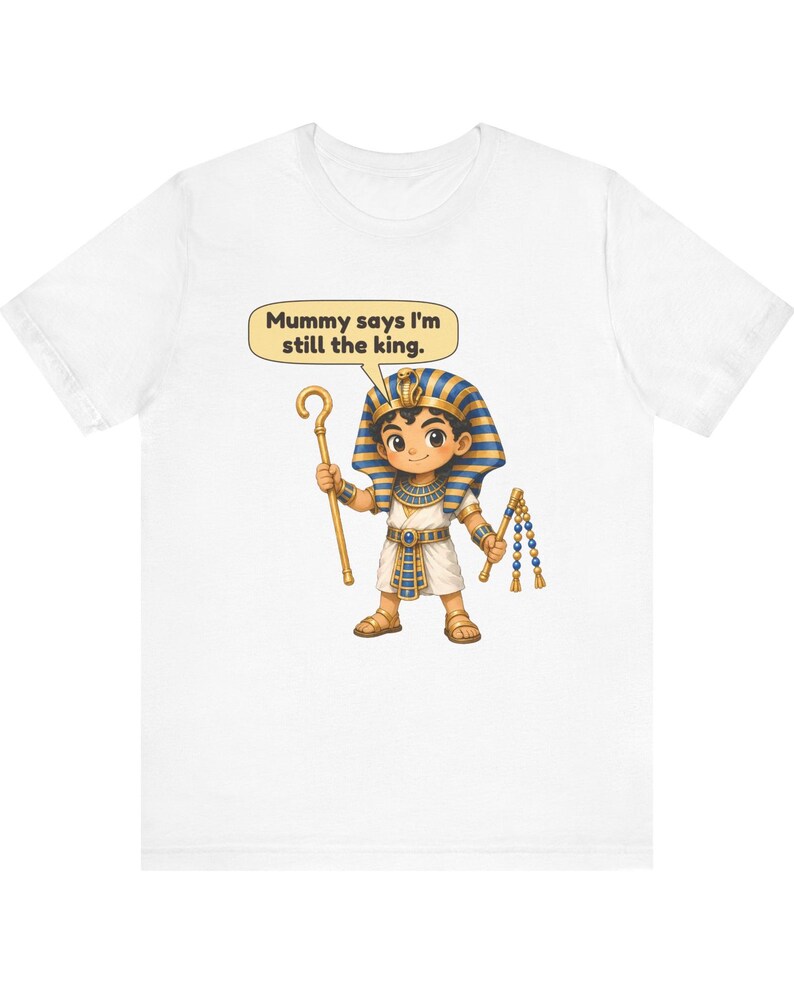 Może przedstawiać: Biały t-shirt z rysunkową ilustracją młodego faraona w niebiesko-złotym nakryciu głowy, trzymającego złote berło i bicz. Dymek nad postacią zawiera tekst, Mummy says I'm still the king.