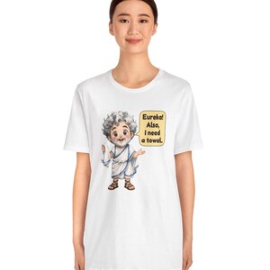 May include: A white t-shirt featuring a cartoon illustration of a bearded man in a white toga holding a white towel. A yellow speech bubble next to the character contains the black text: Eureka! Also, I need a towel.