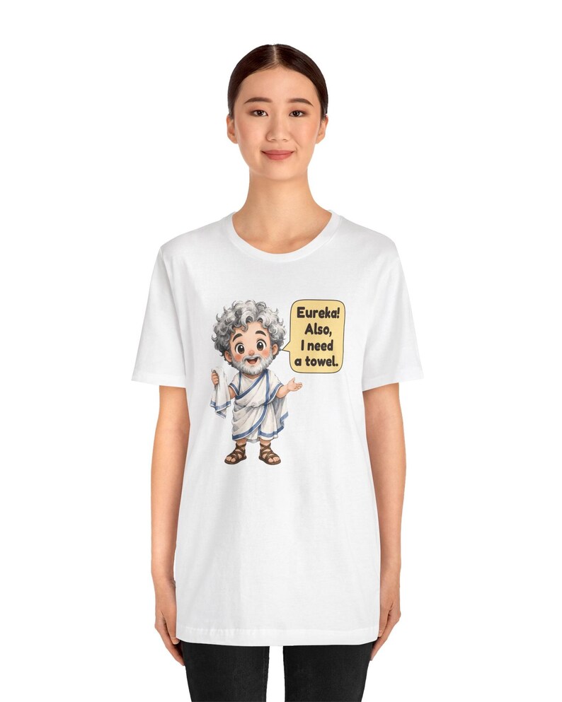 May include: A white t-shirt featuring a cartoon illustration of a bearded man in a white toga holding a white towel. A yellow speech bubble next to the character contains the black text: Eureka! Also, I need a towel.