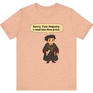 May include: A peach-colored t-shirt featuring a cartoon illustration of a figure in a dark robe, a gold chain, and a halo. A speech bubble above the figure contains the text: Sorry, Your Majesty. I read the fine print.
