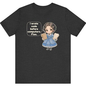 May include: A dark heather gray t-shirt featuring a cartoon illustration of a person in a blue dress holding punch cards and papers. A speech bubble next to the figure contains the text: I wrote code before computers. Flex.