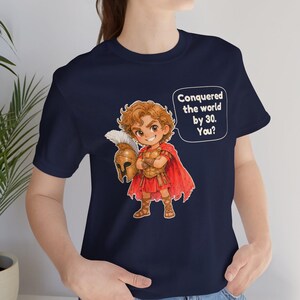 May include: A navy blue t-shirt featuring a cartoon illustration of a historical warrior in golden armor and a red cape, holding a helmet. A speech bubble next to the character contains the white text: Conquered the world by 30. You?