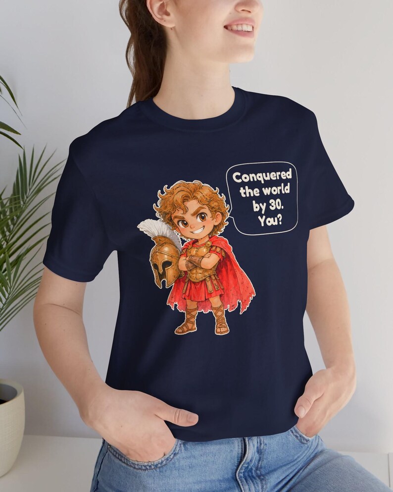 May include: A navy blue t-shirt featuring a cartoon illustration of a historical warrior in golden armor and a red cape, holding a helmet. A speech bubble next to the character contains the white text: Conquered the world by 30. You?
