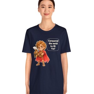 May include: A navy blue t-shirt featuring a cartoon illustration of a warrior in red armor and a cape, holding a plumed helmet. A speech bubble next to the character contains the text: Conquered the world by 30. You?