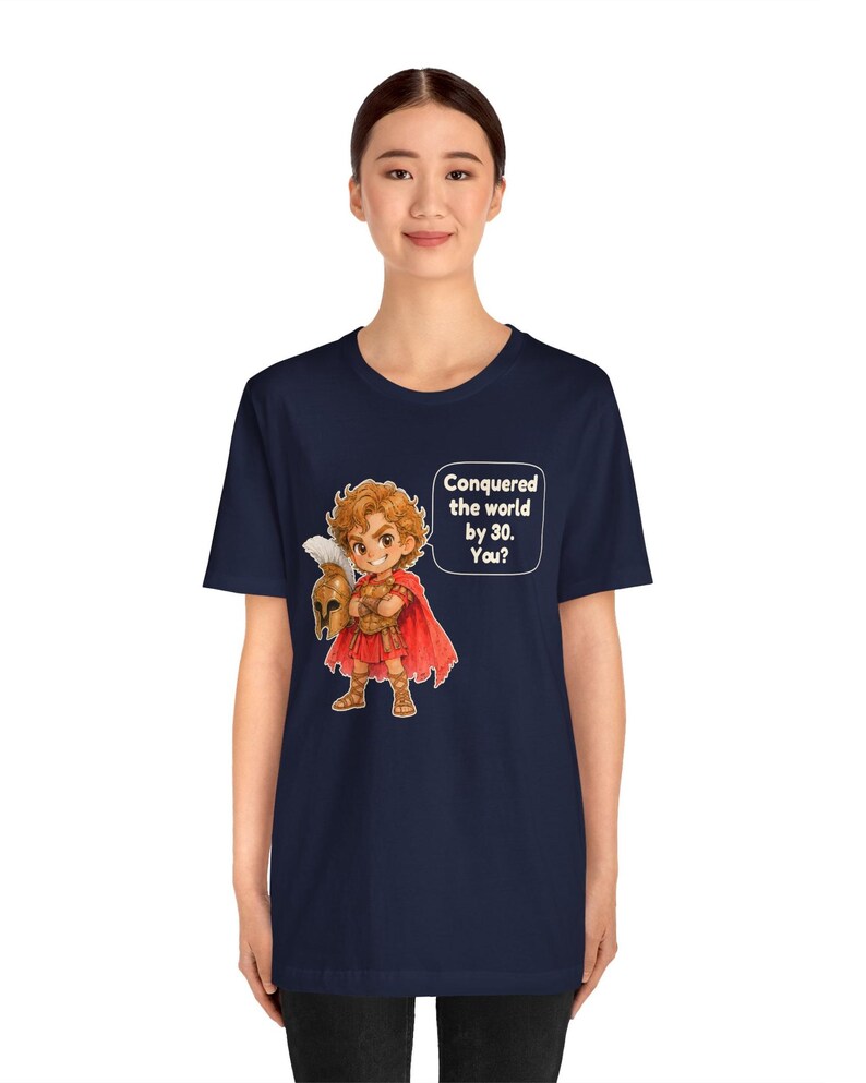 May include: A navy blue t-shirt featuring a cartoon illustration of a warrior in red armor and a cape, holding a plumed helmet. A speech bubble next to the character contains the text: Conquered the world by 30. You?
