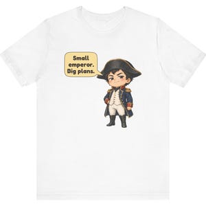 May include: A white t-shirt featuring a graphic of a cartoon character dressed in a historical military uniform with a bicorne hat. Next to the character is a speech bubble containing the text: Small emperor. Big plans.