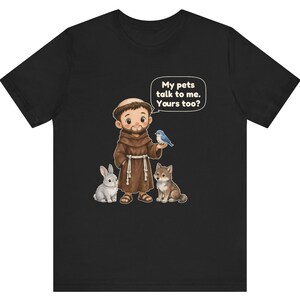 May include: A black short-sleeve t-shirt featuring a cartoon illustration of a monk in a brown robe with a white rope belt. The monk is surrounded by a rabbit, a wolf, and a blue bird. A speech bubble contains the text: My pets talk to me. Yours too?