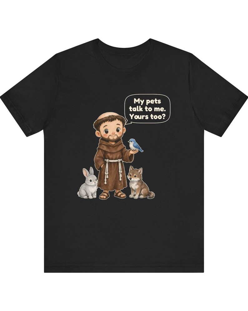 May include: A black short-sleeve t-shirt featuring a cartoon illustration of a monk in a brown robe with a white rope belt. The monk is surrounded by a rabbit, a wolf, and a blue bird. A speech bubble contains the text: My pets talk to me. Yours too?