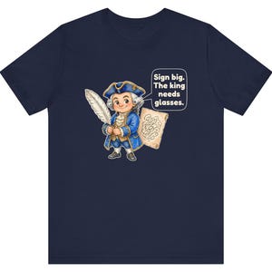 May include: A navy blue t-shirt featuring a cartoon illustration of a person in 18th-century attire holding a large white quill and a scroll. A speech bubble contains the text: Sign big. The king needs glasses.