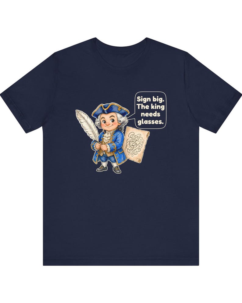 May include: A navy blue t-shirt featuring a cartoon illustration of a person in 18th-century attire holding a large white quill and a scroll. A speech bubble contains the text: Sign big. The king needs glasses.