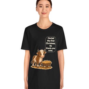 以下が含まれることがあります： 黒の半袖Tシャツ。干し草が入った木製の飼い葉桶の横に立つ、茶色と白の子牛のグラフィックが描かれています。子牛の上の吹き出しには、白い文字で「Hosted the first Christmas. No thank-you note.」と書かれています。