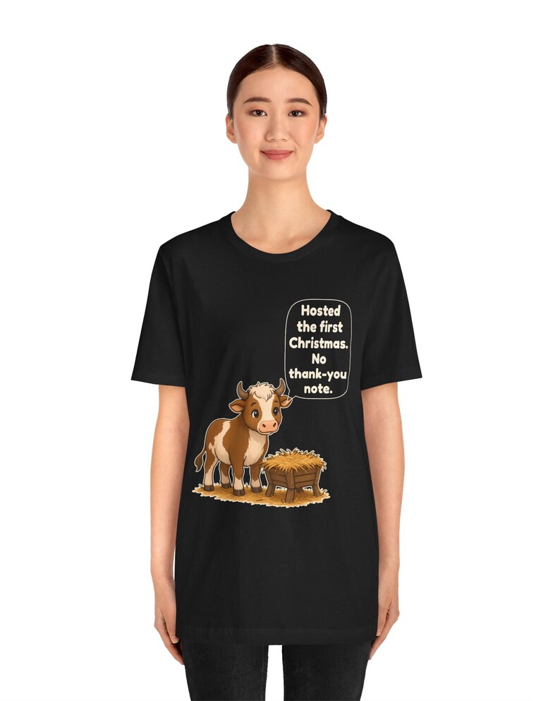 以下が含まれることがあります： 黒の半袖Tシャツ。干し草が入った木製の飼い葉桶の横に立つ、茶色と白の子牛のグラフィックが描かれています。子牛の上の吹き出しには、白い文字で「Hosted the first Christmas. No thank-you note.」と書かれています。
