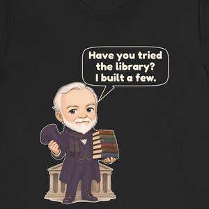 May include: A black t-shirt featuring a cartoon illustration of an older man with a white beard, wearing a dark coat and scarf, holding a top hat and a stack of books. A speech bubble contains the text: Have you tried the library? I built a few.