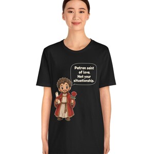 Puede incluir: Una camiseta negra con una ilustraci&oacute;n de dibujos animados de una figura barbuda con t&uacute;nica roja que sostiene un pergamino y una rosa roja. Un globo de texto sobre la figura contiene el texto: Patron saint of love. Not your situationship.