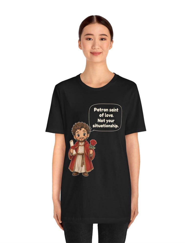 Puede incluir: Una camiseta negra con una ilustraci&oacute;n de dibujos animados de una figura barbuda con t&uacute;nica roja que sostiene un pergamino y una rosa roja. Un globo de texto sobre la figura contiene el texto: Patron saint of love. Not your situationship.