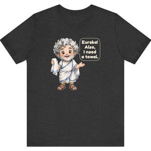 May include: A dark heather gray t-shirt featuring a cartoon illustration of a bearded figure in a white toga holding a towel. A speech bubble next to the figure contains the text: Eureka! Also, I need a towel. The design is centered on the chest.