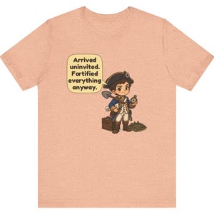 May include: A peach-colored t-shirt featuring a cartoon illustration of a historical explorer in a blue coat and tricorn hat, holding a shovel and compass, with a speech bubble that reads: Arrived uninvited. Fortified everything anyway.