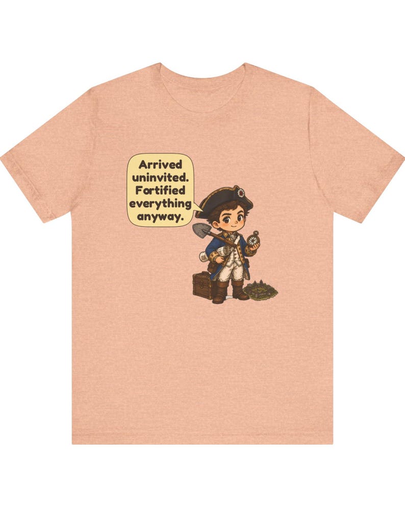 May include: A peach-colored t-shirt featuring a cartoon illustration of a historical explorer in a blue coat and tricorn hat, holding a shovel and compass, with a speech bubble that reads: Arrived uninvited. Fortified everything anyway.