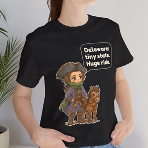 May include: A black short-sleeve t-shirt featuring a cartoon illustration of a person in a tricorn hat and colonial-style coat standing next to a small brown horse. A speech bubble contains the text: Delaware: tiny state. Huge ride.
