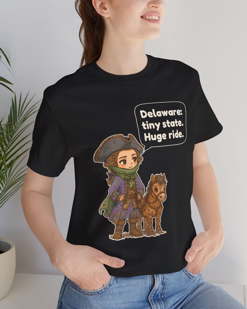 May include: A black short-sleeve t-shirt featuring a cartoon illustration of a person in a tricorn hat and colonial-style coat standing next to a small brown horse. A speech bubble contains the text: Delaware: tiny state. Huge ride.