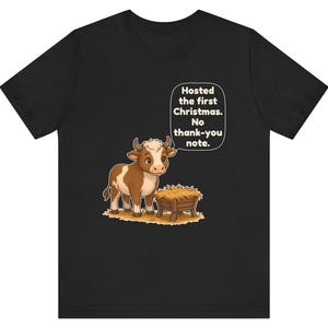 以下が含まれることがあります： 黒の半袖Tシャツ。干し草が入った木製の飼い葉桶の横に立つ茶色と白の子牛のグラフィックが描かれています。子牛の上の吹き出しには「Hosted the first Christmas. No thank-you note.」というテキストが含まれています。