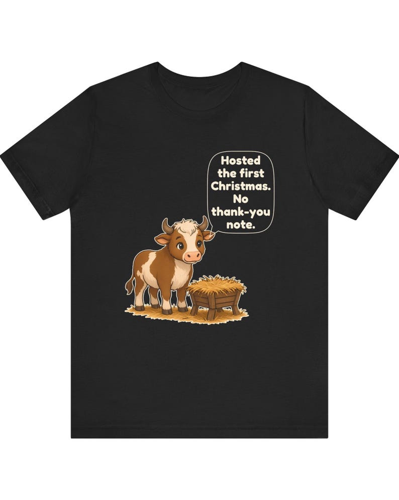 以下が含まれることがあります： 黒の半袖Tシャツ。干し草が入った木製の飼い葉桶の横に立つ茶色と白の子牛のグラフィックが描かれています。子牛の上の吹き出しには「Hosted the first Christmas. No thank-you note.」というテキストが含まれています。